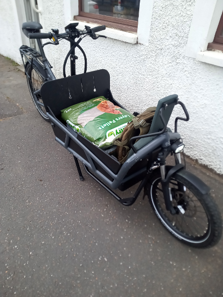 A grey cargo bike parked on pavement beside the white wall of a buiding